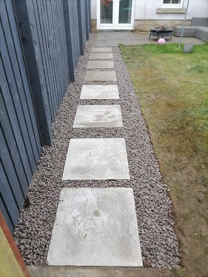 A path of grey slabs set in gravel beside a dark fence